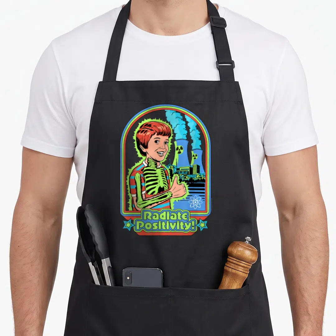 Radiate steven rhodes apron - Boy with green skeleton giving thumbs up near nuclear towers with text Radiate Positivity — shown on a black cotton canvas apron against a white background