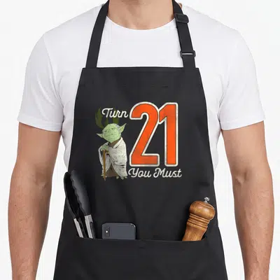 Yoda 21st birthday apron - Cartoon Yoda standing beside orange number 21 with white text Turn You Must — shown on a black cotton canvas apron against a white background