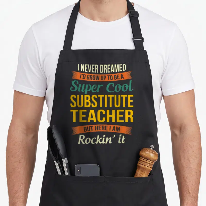 Cute substitute teacher apron - Stacked text reading I NEVER DREAMED I'D GROW UP TO BE A Super Cool SUBSTITUTE TEACHER — shown on a black cotton canvas apron against a white background