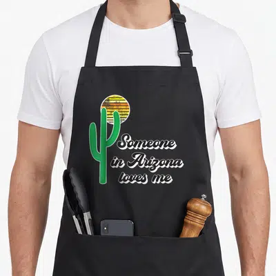 Funny someone arizona apron - Green saguaro cactus next to striped sun and black cursive text saying Someone in Arizona — shown on a black cotton canvas apron against a white background
