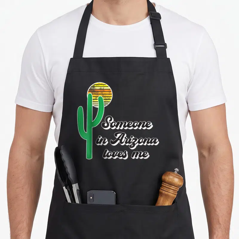 Funny someone arizona apron - Green saguaro cactus next to striped sun and black cursive text saying Someone in Arizona — shown on a black cotton canvas apron against a white background