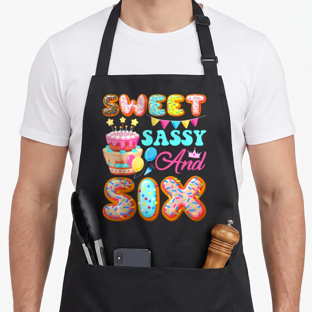 Six sassy apron - Donut letters spelling Sweet Sassy And Six with cake and bunting — shown on a black cotton canvas apron against a white background