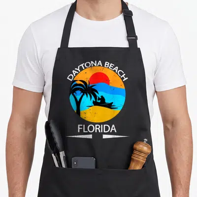 Cute daytona florida apron - Retro circular graphic with sun, black palm tree, jet ski, and DAYTONA BEACH FLORIDA text — shown on a black cotton canvas apron against a white background