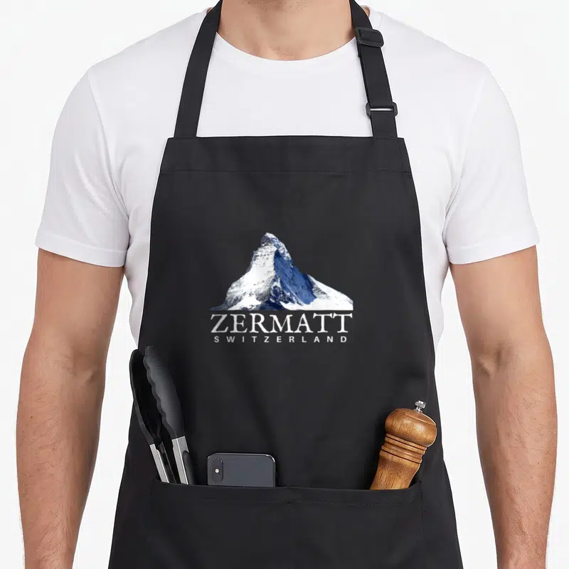 Zermatt swiss apron - Snow-capped mountain peak photo above stacked white text ZERMATT SWITZERLAND — shown on a black cotton canvas apron against a white background