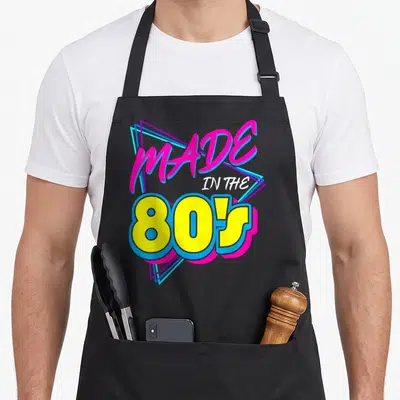 Nineteen 80s apron - Retro text graphic MADE IN THE 80S with pink script and yellow block letters over nested triangles — shown on a black cotton canvas apron against a white background