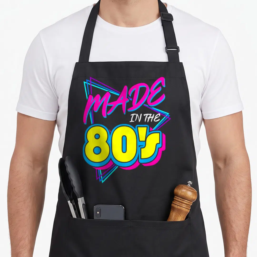 Nineteen 80s apron - Retro text graphic MADE IN THE 80S with pink script and yellow block letters over nested triangles — shown on a black cotton canvas apron against a white background