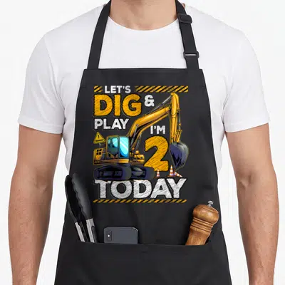 Construction 2nd apron - Yellow excavator with LET'S DIG & PLAY I'M 2 TODAY text and traffic cones — shown on a black cotton canvas apron against a white background
