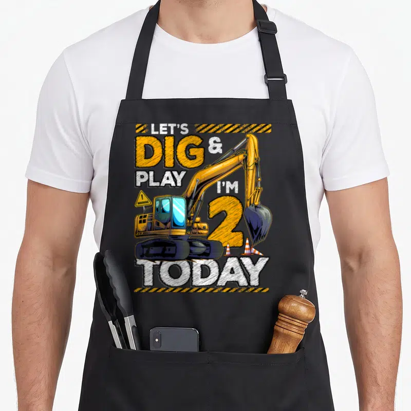 Construction 2nd apron - Yellow excavator with LET'S DIG & PLAY I'M 2 TODAY text and traffic cones — shown on a black cotton canvas apron against a white background