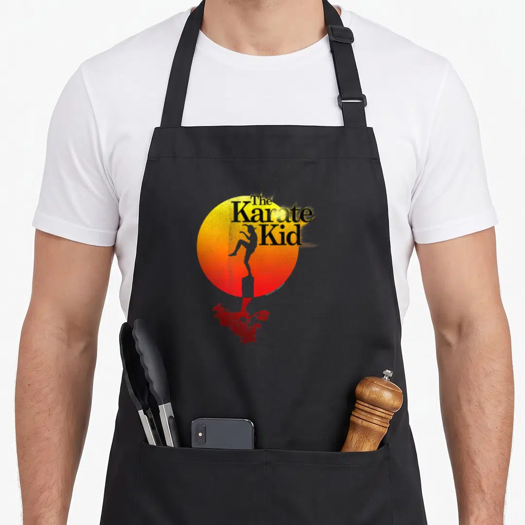 Sun karate apron - Retro sun graphic with The Karate Kid text and silhouette standing on a post above a red tree — shown on a black cotton canvas apron against a white background