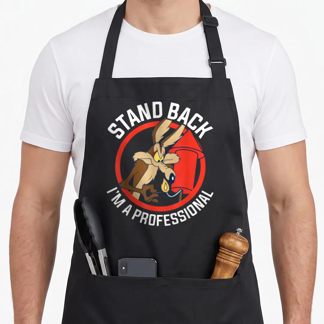 Coyote professional aprons - Wile E. Coyote holding a lit red rocket inside a circle with white text reading STAND BACK — shown on a black cotton canvas apron against a white background