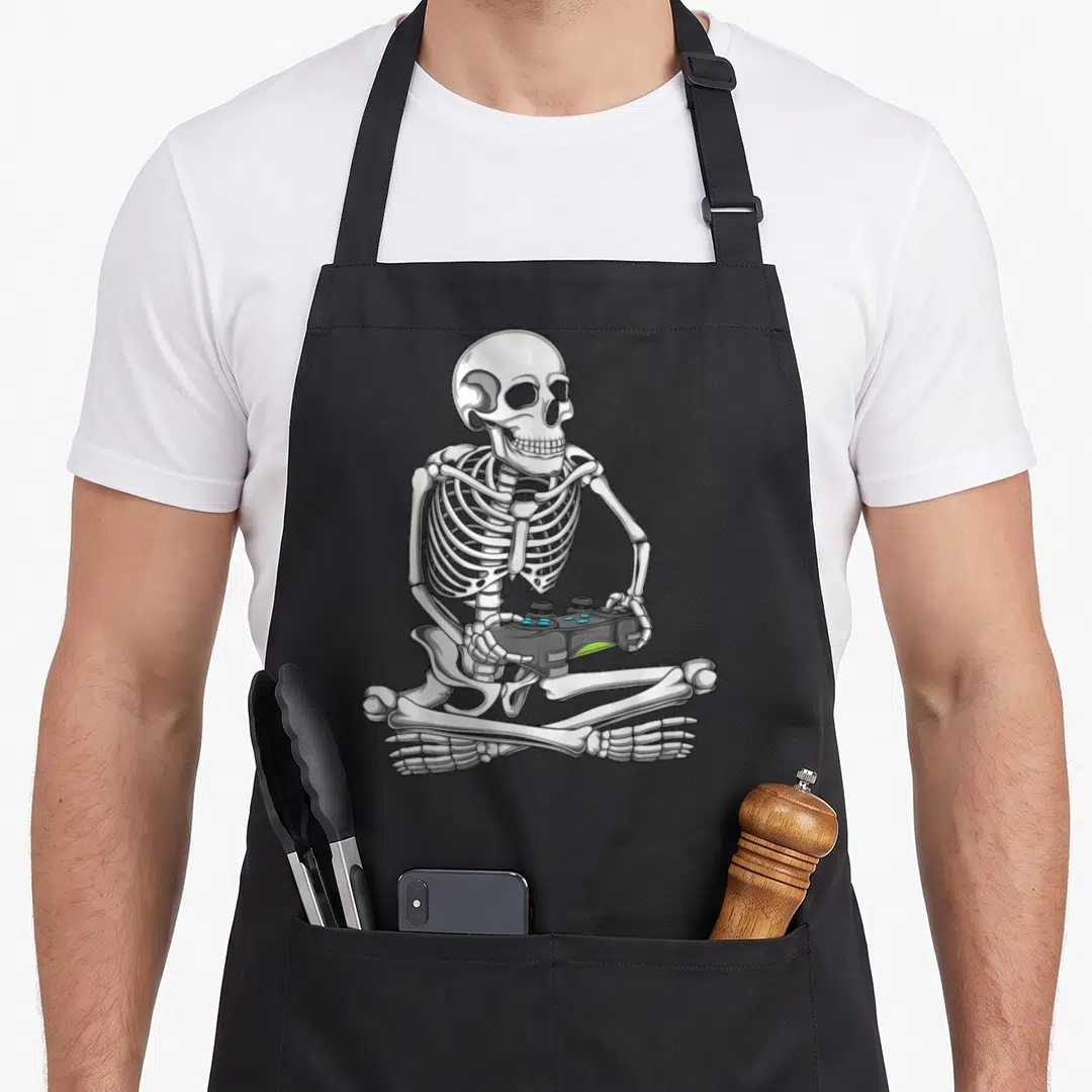 Skeleton gaming halloween apron - Cartoon skeleton sitting cross-legged holding a black game controller in white — shown on a black cotton canvas apron against a white background