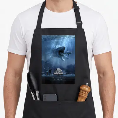 Mosasaurus jurassic world apron - Blue underwater poster with dinosaur eating shark and text Jurassic World The Park Is — shown on a black cotton canvas apron against a white background