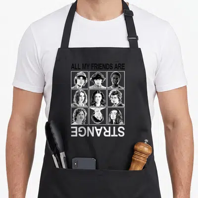 Strange friends cooking apron - Nine retro portraits in white frames with text ALL MY FRIENDS ARE STRANGE upside down — shown on a black cotton canvas apron against a white background