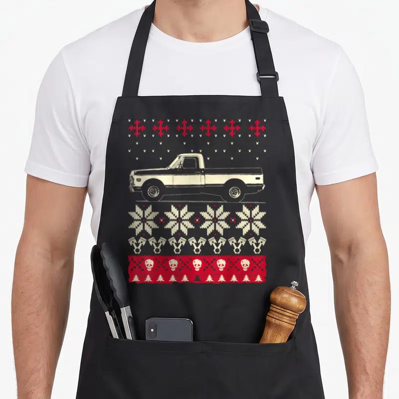 Pickup ugly christmas sweater apron - Beige pickup truck with red snowflakes, beige pistons, skulls, and pine tree rows — shown on a black cotton canvas apron against a white background