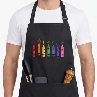 Mood teacher apron - Seven cartoon crayons in rainbow hues with faces and floating emotion symbols like sun, hearts — shown on a black cotton canvas apron against a white background