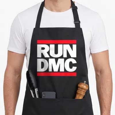 Run novelty apron - White RUN DMC text between two red horizontal bars — shown on a black cotton canvas apron against a white background