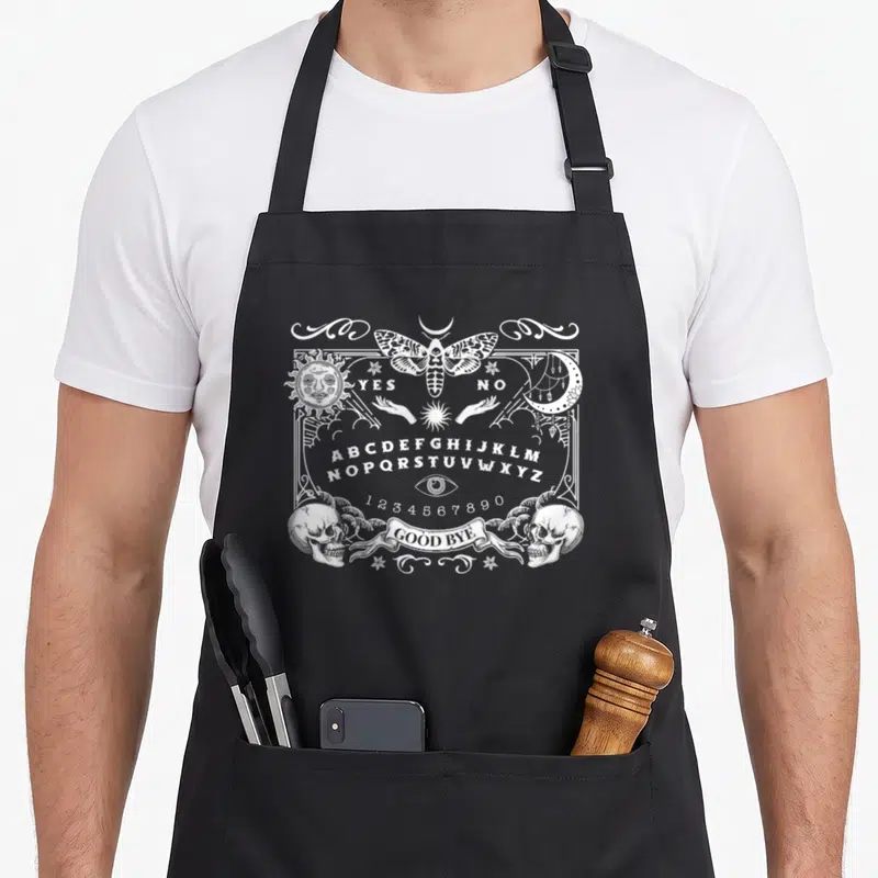 Board halloween apron - White line art ouija board with moth, sun, moon, skulls, and GOOD BYE banner — shown on a black cotton canvas apron against a white background