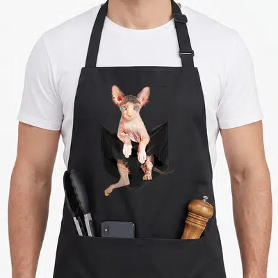 Cute sphynx cat apron - Realistic pinkish-beige sphynx cat emerging from black pocket with torn bottom holes — shown on a black cotton canvas apron against a white background