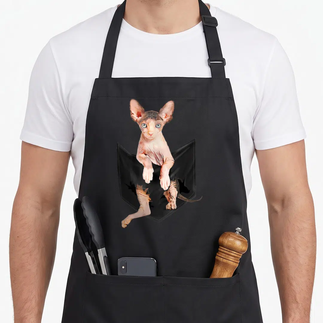 Cute sphynx cat apron - Realistic pinkish-beige sphynx cat emerging from black pocket with torn bottom holes — shown on a black cotton canvas apron against a white background