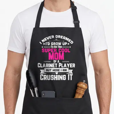 Player pipe mom apron - Stacked white and pink text reading Super Cool Mom Of A Clarinet Player with decorative stars — shown on a black cotton canvas apron against a white background