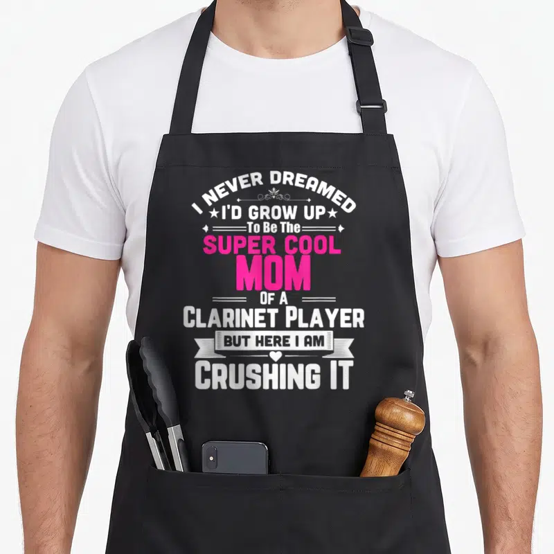 Player pipe mom apron - Stacked white and pink text reading Super Cool Mom Of A Clarinet Player with decorative stars — shown on a black cotton canvas apron against a white background