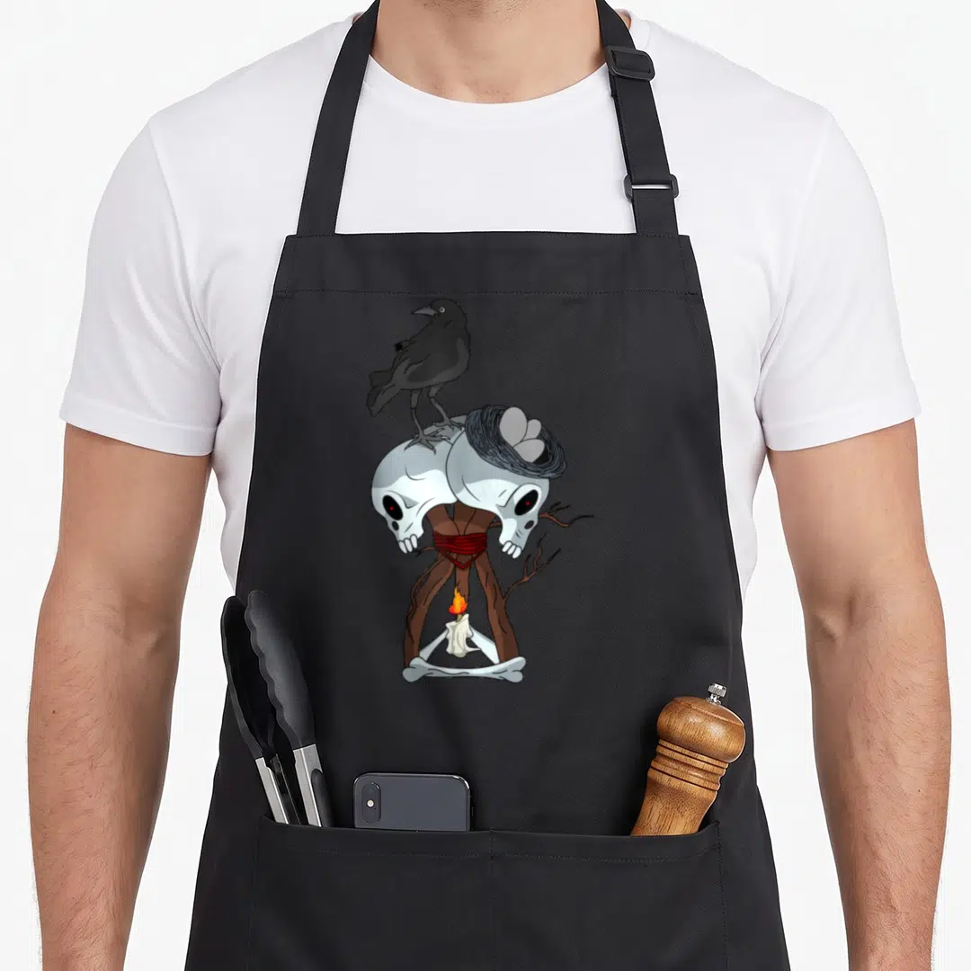 Skull video game apron - Cartoon crow on two skulls with nest and candle between crossed bones — shown on a black cotton canvas apron against a white background