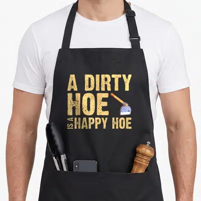 Dirty gardener apron - Distressed text A DIRTY HOE IS A HAPPY HOE with an orange handle blue blade hoe graphic — shown on a black cotton canvas apron against a white background