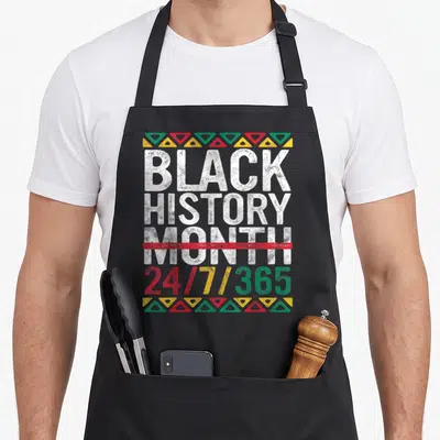 Pride african print apron - Stacked text Black History Month 24/7/365 with red strikethrough and triangle borders — shown on a black cotton canvas apron against a white background