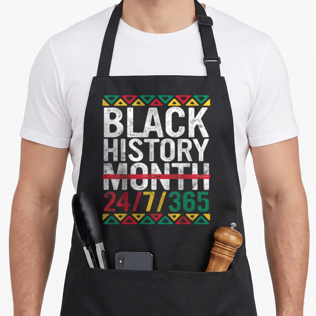 Pride african print apron - Stacked text Black History Month 24/7/365 with red strikethrough and triangle borders — shown on a black cotton canvas apron against a white background