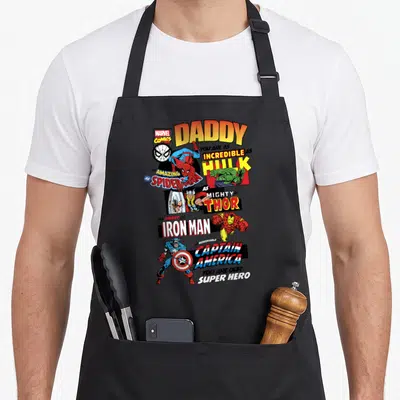 Comic father day apron - Vertical stack of cartoon superheroes with DADDY text in orange, yellow, red, blue, green — shown on a black cotton canvas apron against a white background
