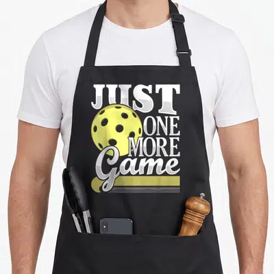 Game player pickleball apron - Yellow perforated ball next to stacked white JUST ONE MORE Game text on gray — shown on a black cotton canvas apron against a white background