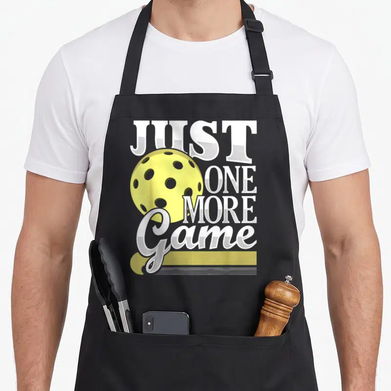 Game player pickleball apron - Yellow perforated ball next to stacked white JUST ONE MORE Game text on gray — shown on a black cotton canvas apron against a white background