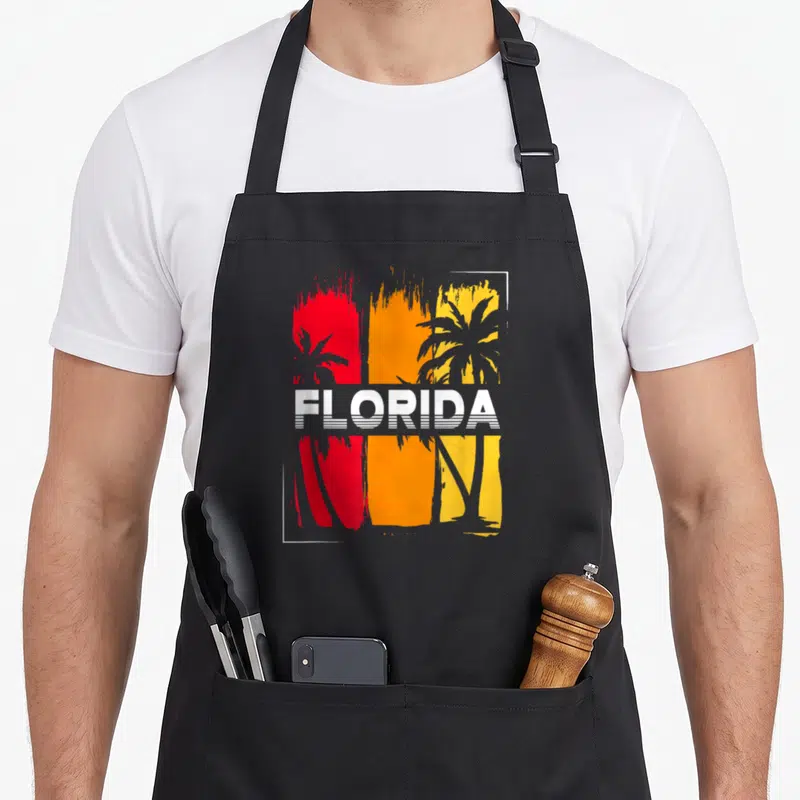 South summer aprons - Retro Florida text over red orange yellow stripes with black palm trees — shown on a black cotton canvas apron against a white background