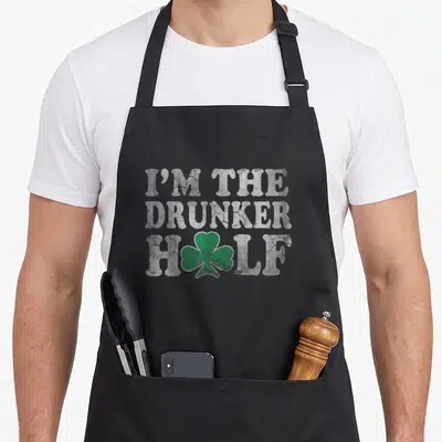 Drunker st patrick day aprons - I'M THE DRUNKER HALF text with green shamrock replacing A in distressed white font — shown on a black cotton canvas apron against a white background