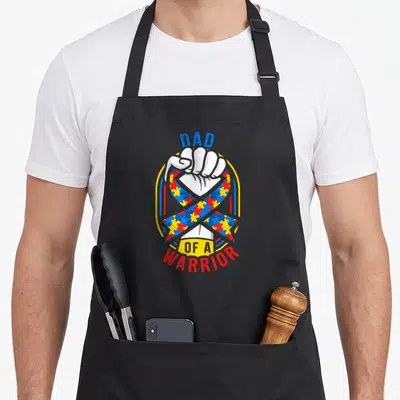 Month autism dad apron - Dad of a Warrior text surrounding a fist holding a colorful puzzle piece ribbon — shown on a black cotton canvas apron against a white background