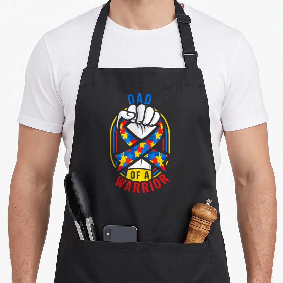 Month autism dad apron - Dad of a Warrior text surrounding a fist holding a colorful puzzle piece ribbon — shown on a black cotton canvas apron against a white background