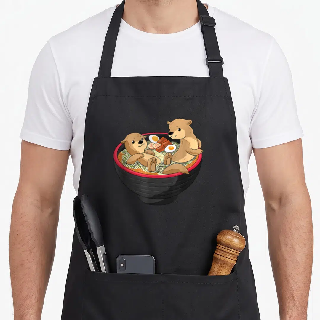Otter kawaii apron - Two cartoon otters in a black ramen bowl with eggs and meat in brown, black, red, yellow — shown on a black cotton canvas apron against a white background