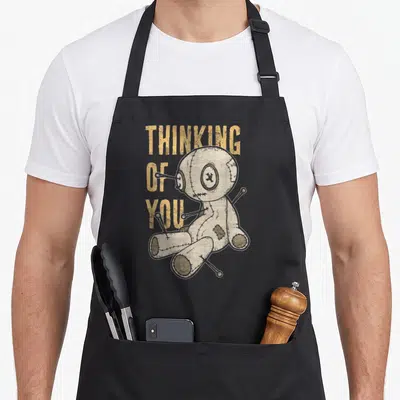 Thinking goth apron - Voodoo doll with pins next to three stacked words THINKING OF YOU in yellow and beige — shown on a black cotton canvas apron against a white background