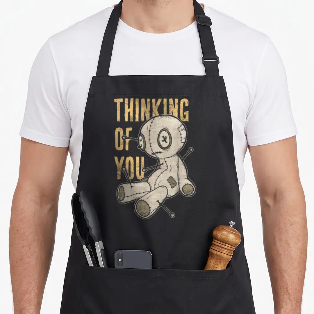 Thinking goth apron - Voodoo doll with pins next to three stacked words THINKING OF YOU in yellow and beige — shown on a black cotton canvas apron against a white background