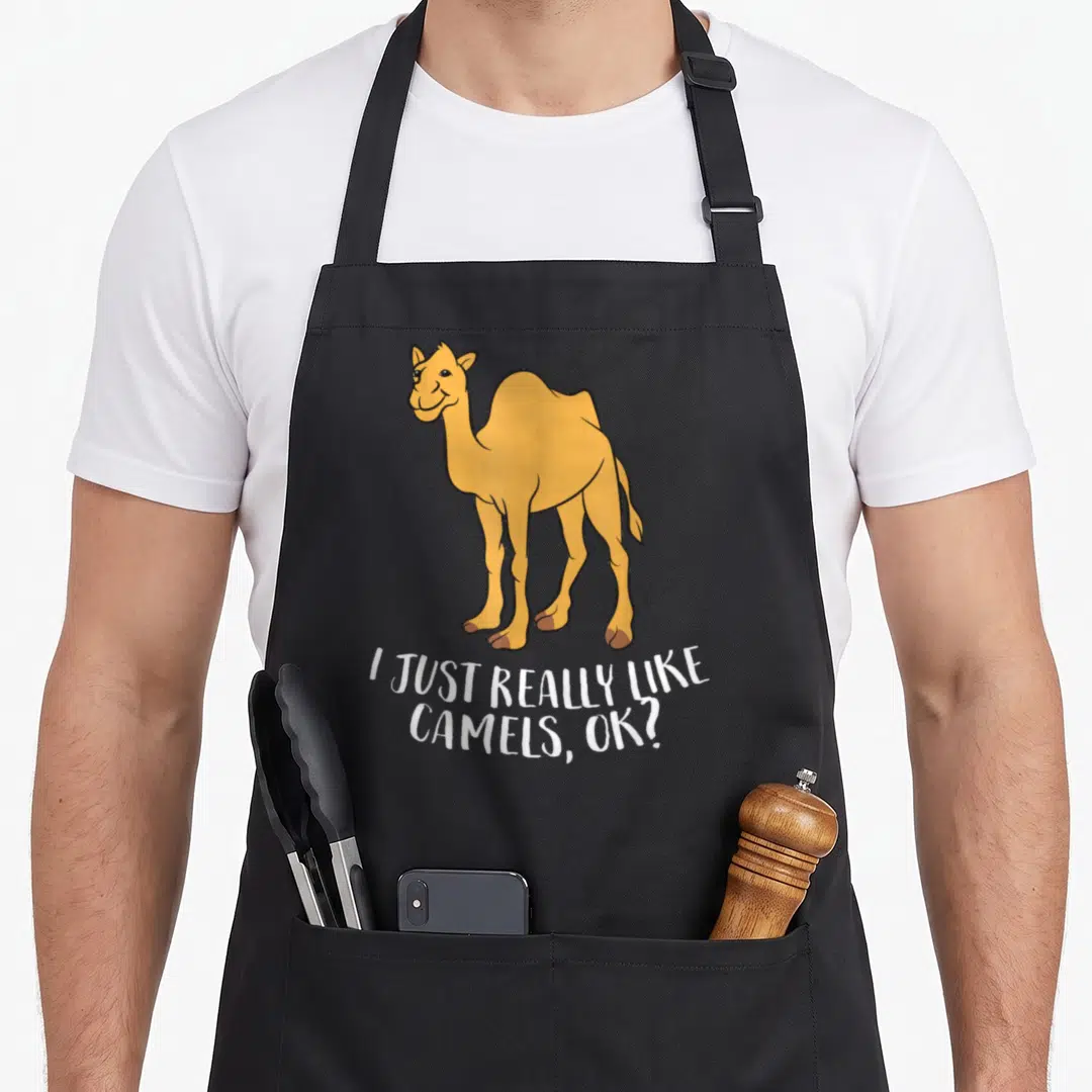 Funny camel apron - Cartoon yellow-orange camel standing above white text reading I JUST REALLY LIKE CAMELS, OK? — shown on a black cotton canvas apron against a white background