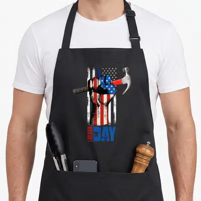 Fist american flag apron - Fist holding hammer over distressed red white blue flag with LABOUR DAY text and wheelbarrow — shown on a black cotton canvas apron against a white background