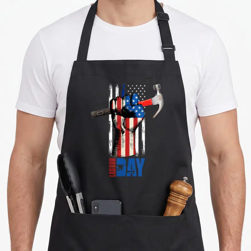 Fist american flag apron - Fist holding hammer over distressed red white blue flag with LABOUR DAY text and wheelbarrow — shown on a black cotton canvas apron against a white background
