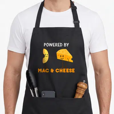 Powered cheese mac apron - Cartoon macaroni noodle and cheese wedge standing side by side with POWERED BY MAC & CHEESE — shown on a black cotton canvas apron against a white background