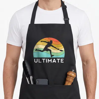 Flying ultimate apron - Retro player silhouette throwing disc under multicolor sunset with ULTIMATE text — shown on a black cotton canvas apron against a white background