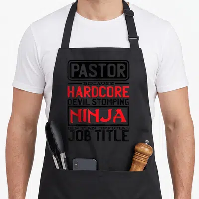 Prist devil's apron - Stacked typography text Pastor Because Hardcore Devil Stomping Ninja Isnt Official Job Title — shown on a black cotton canvas apron against a white background