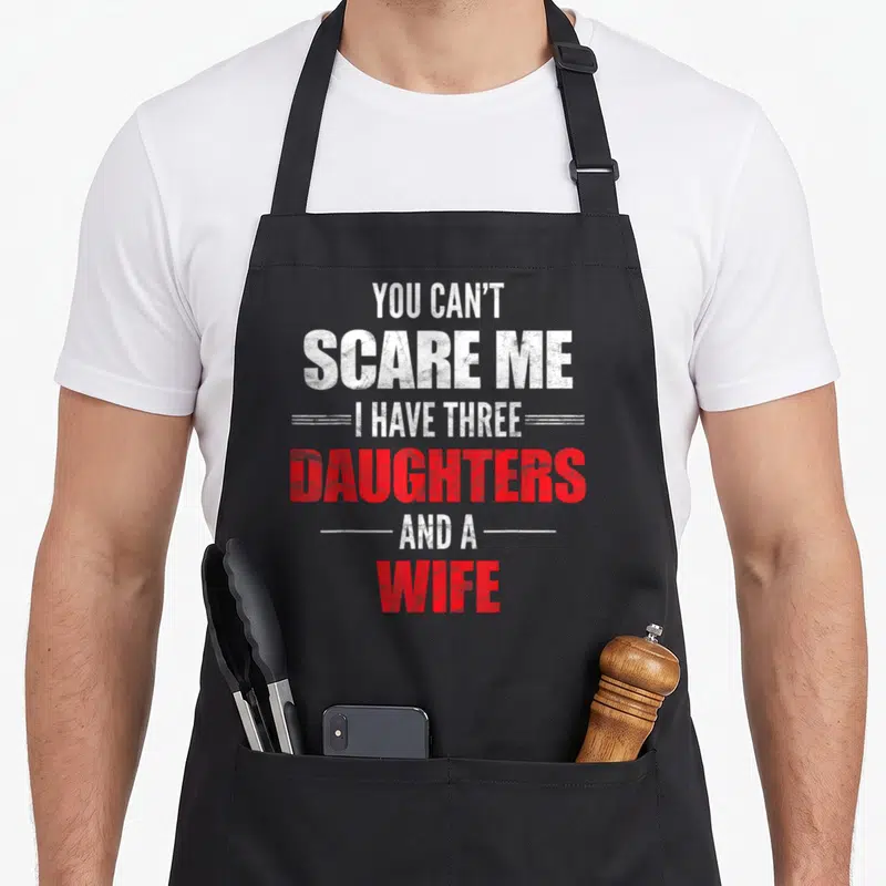 Three wife apron - Distressed white and red text reading You Can't Scare Me I Have Three Daughters And A Wife — shown on a black cotton canvas apron against a white background