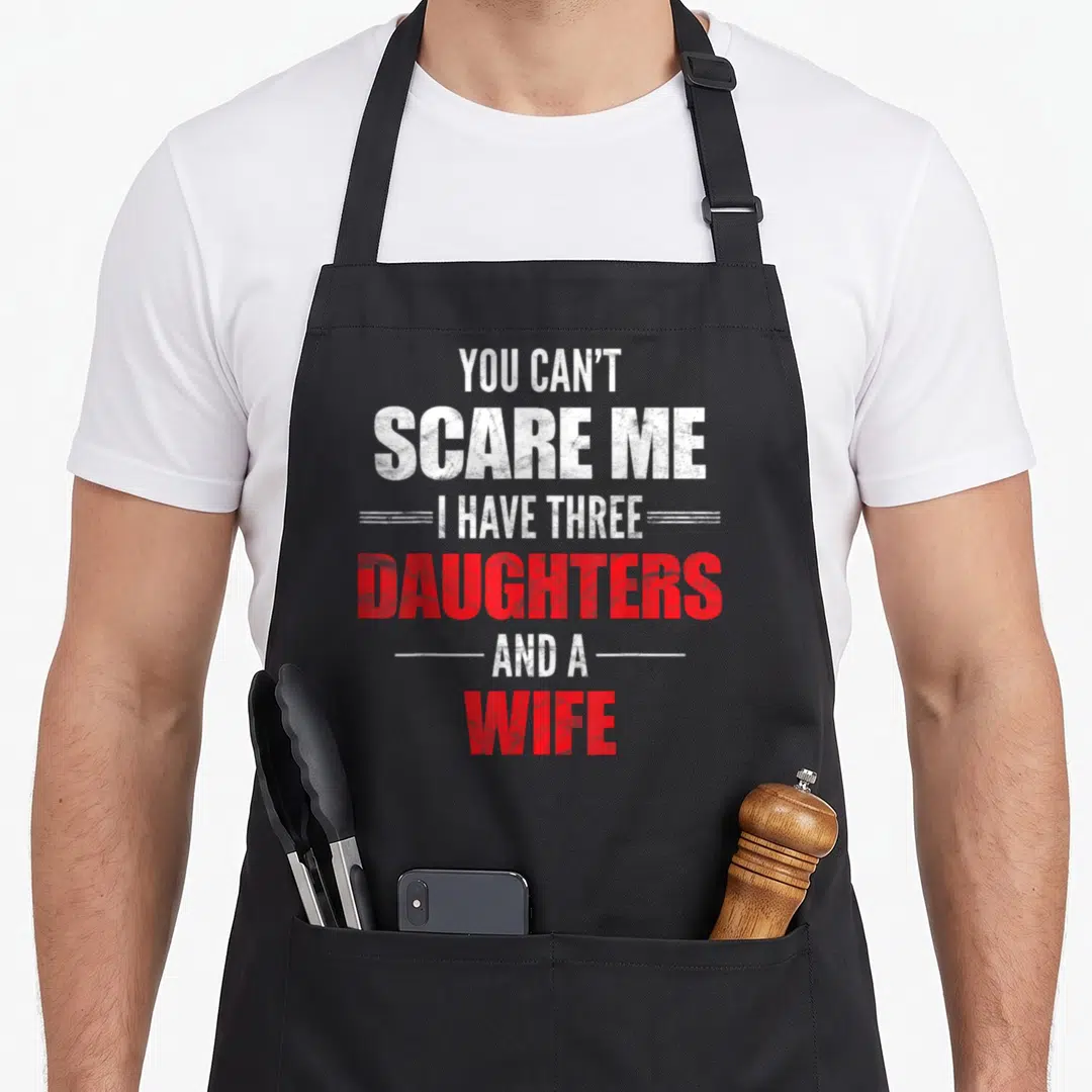 Three wife apron - Distressed white and red text reading You Can't Scare Me I Have Three Daughters And A Wife — shown on a black cotton canvas apron against a white background
