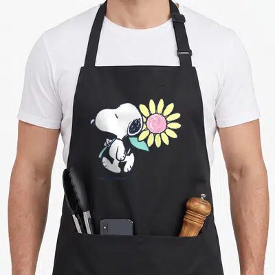 Flower daisy apron - White cartoon dog standing holding a yellow sunflower with a pink center and light blue leaf — shown on a black cotton canvas apron against a white background