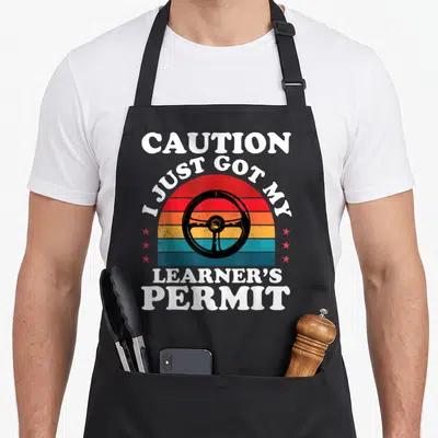 Learner driver permit apron - Retro striped sun with black steering wheel and white text reading CAUTION I JUST GOT MY — shown on a black cotton canvas apron against a white background