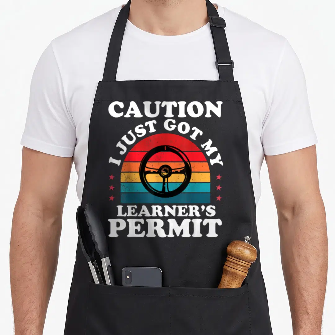 Learner driver permit apron - Retro striped sun with black steering wheel and white text reading CAUTION I JUST GOT MY — shown on a black cotton canvas apron against a white background